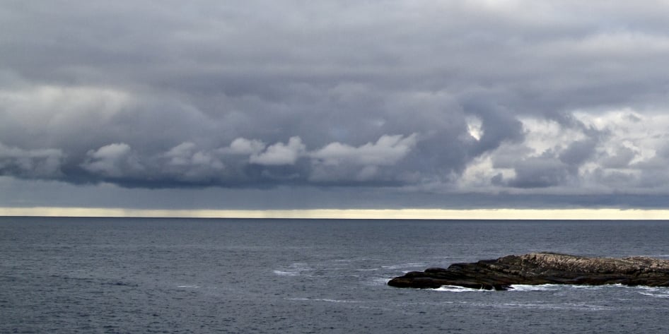 Mørke stormskyger som samler seg over et rolig hav ved en klippeformasjon.